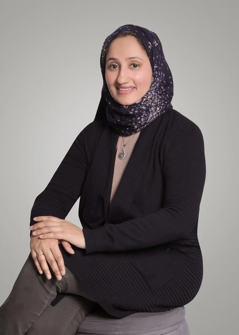 Dr. Rifat Mazhar Falak, smiling, seated, in a black cardigan and navy floral headscarf